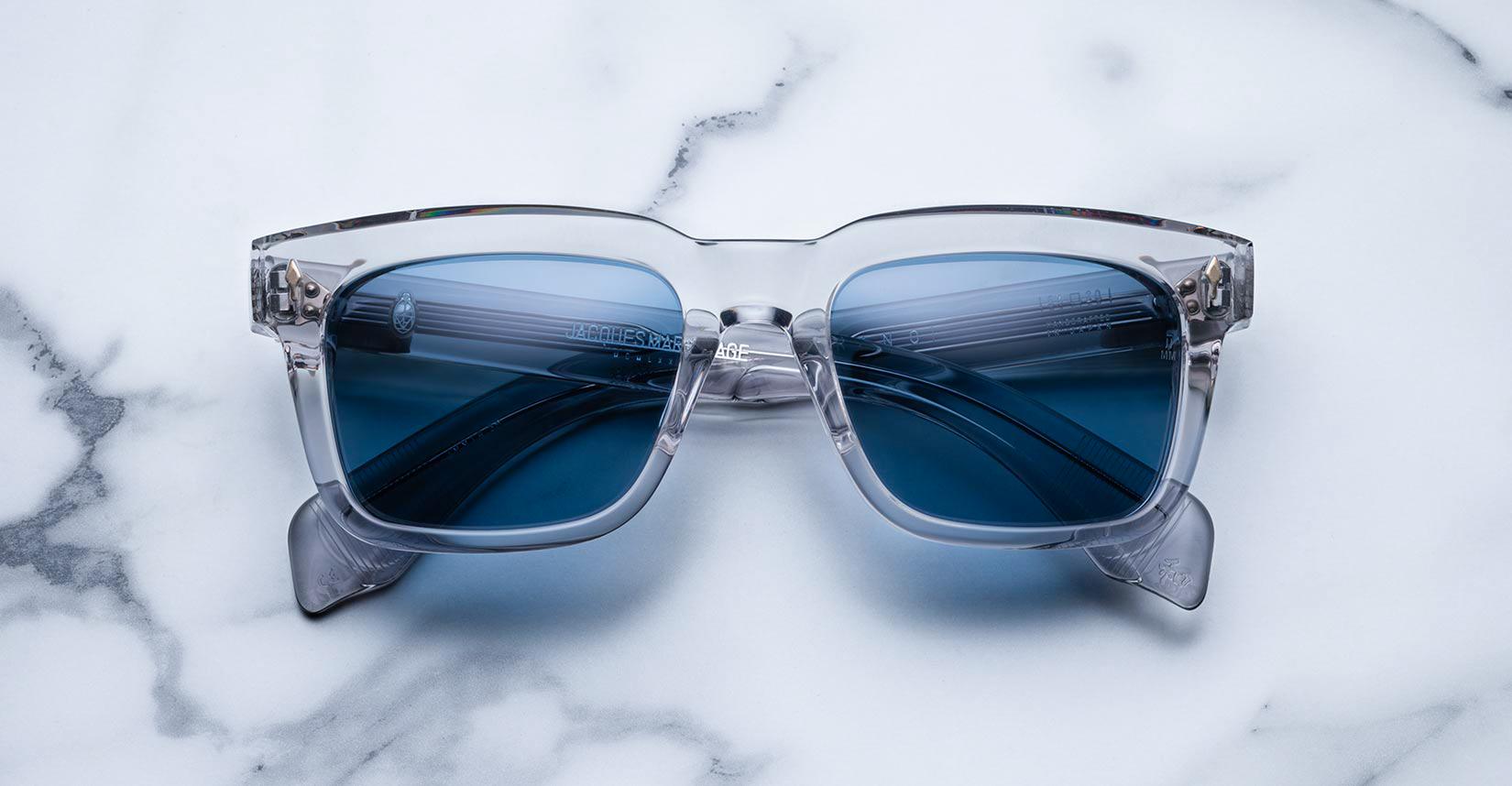 Jacques Marie Mage Torino sunglasses feature clear frames and blue-tinted lenses, inspired by the brand's bold rectangular design, displayed on a white marble surface.
