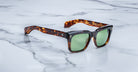 The Jacques Marie Mage Torino handcrafted acetate sunglasses in tortoiseshell with green lenses are displayed on a white marble surface.
