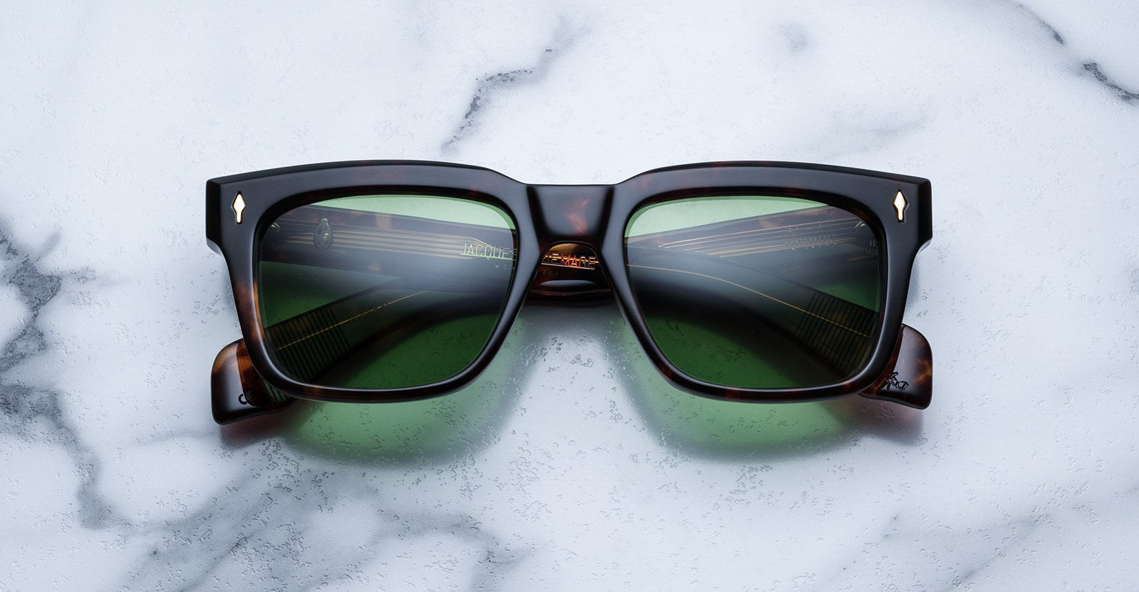 The Jacques Marie Mage Torino eyeglass frames feature bold rectangular tortoiseshell acetate and green lenses, handcrafted by Jacques Marie Mage, shown on a white marble surface.