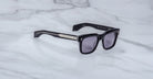 The Jacques Marie Mage Torino by Jacques Marie Mage features bold, handcrafted black acetate rectangular frames with dark lenses and silver arm detailing, displayed on a light gray marble surface.