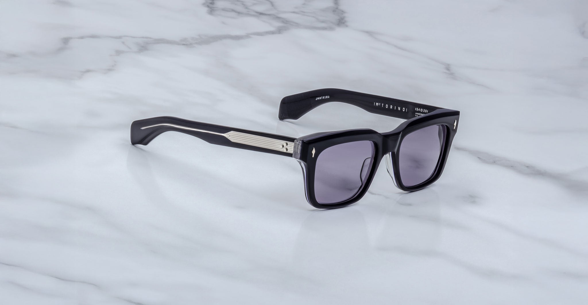 The Jacques Marie Mage Torino by Jacques Marie Mage features bold, handcrafted black acetate rectangular frames with dark lenses and silver arm detailing, displayed on a light gray marble surface.