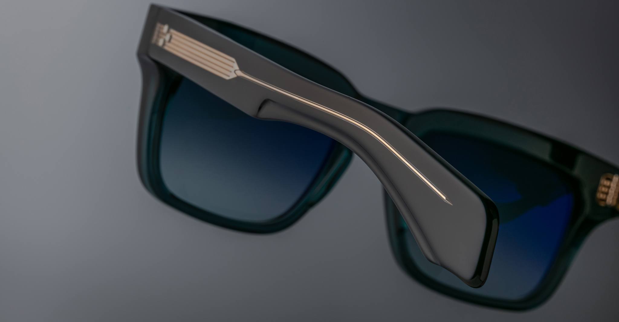 Close-up of Jacques Marie Mage Torino eyewear: bold rectangular frames with blue lenses, highlighting handcrafted acetate design details on the arm and hinge, set against a gray background.