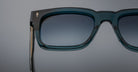 Close-up of Jacques Marie Mage Torino eyeglasses: bold, handcrafted acetate frames in teal with gradient-tinted lenses by Jacques Marie Mage, set against a plain gray background.
