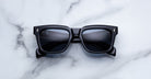 Jacques Marie Mage Torino bold black rectangular eyeglasses by Jacques Marie Mage, featuring dark lenses, are displayed on a white marble surface.