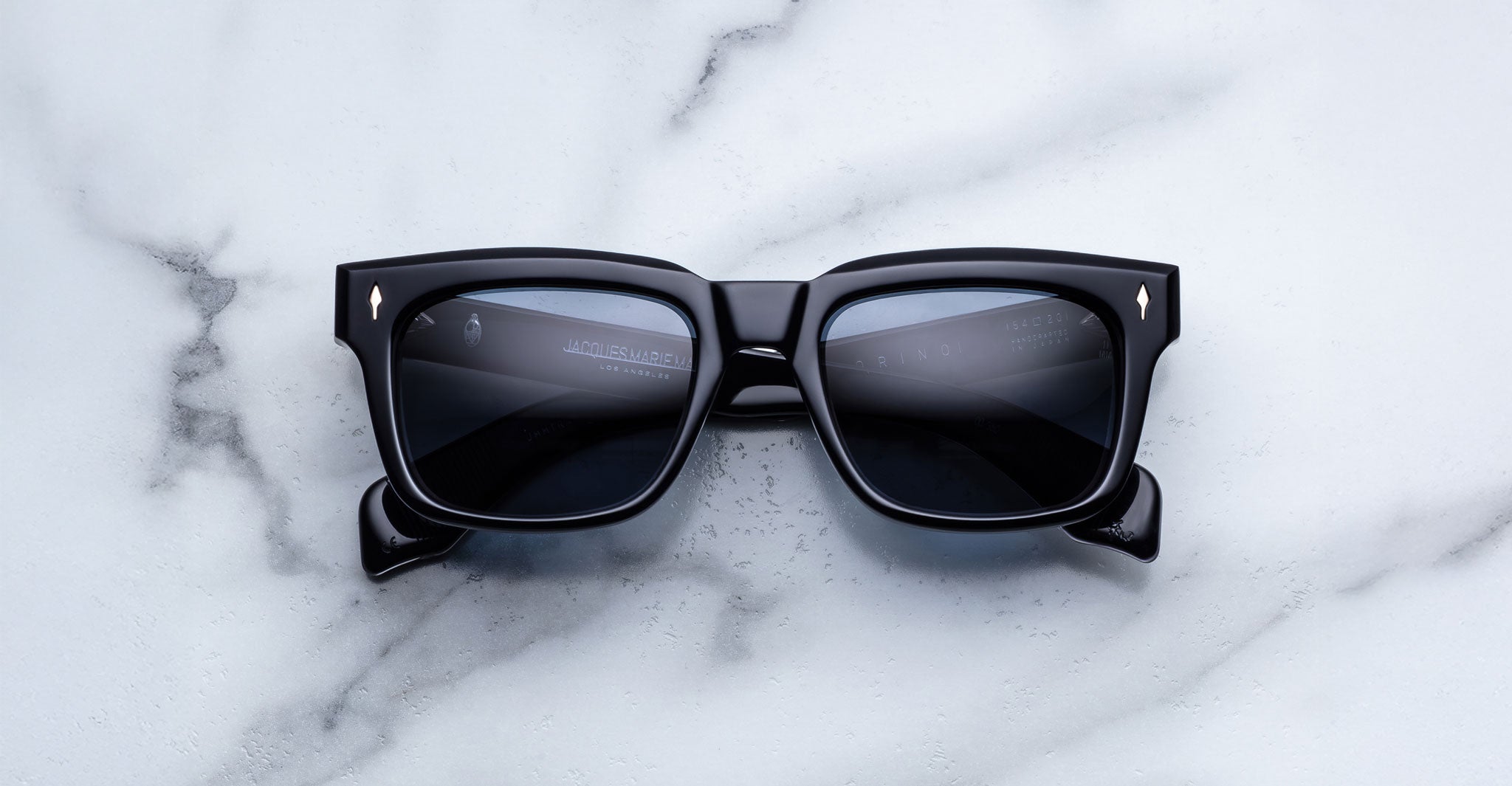 Jacques Marie Mage Torino bold black rectangular eyeglasses by Jacques Marie Mage, featuring dark lenses, are displayed on a white marble surface.