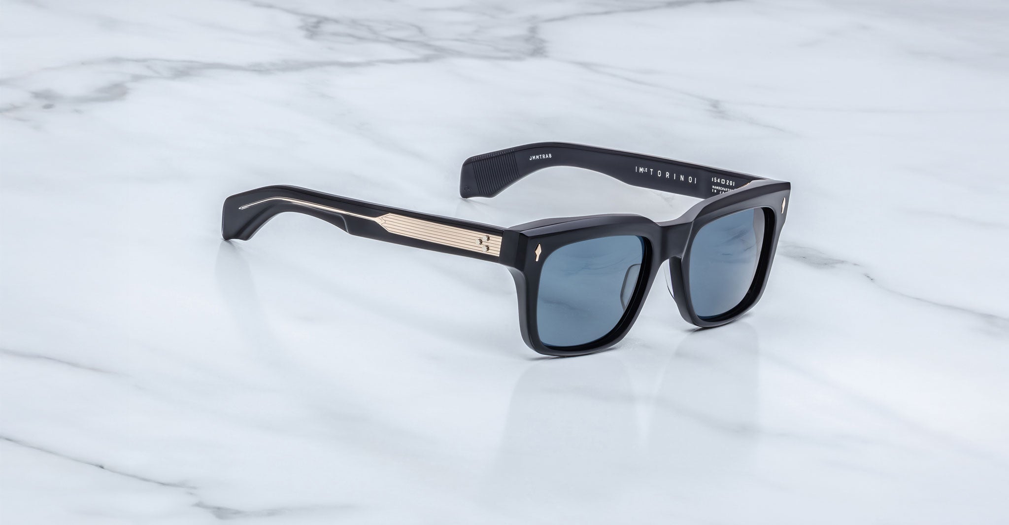 The Jacques Marie Mage Torino by Jacques Marie Mage features black handcrafted acetate rectangular frames with blue-tinted lenses and gold arm detailing, displayed on a white marble surface.