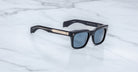 The Jacques Marie Mage Torino by Jacques Marie Mage features black handcrafted acetate rectangular frames with blue-tinted lenses and gold arm detailing, displayed on a white marble surface.