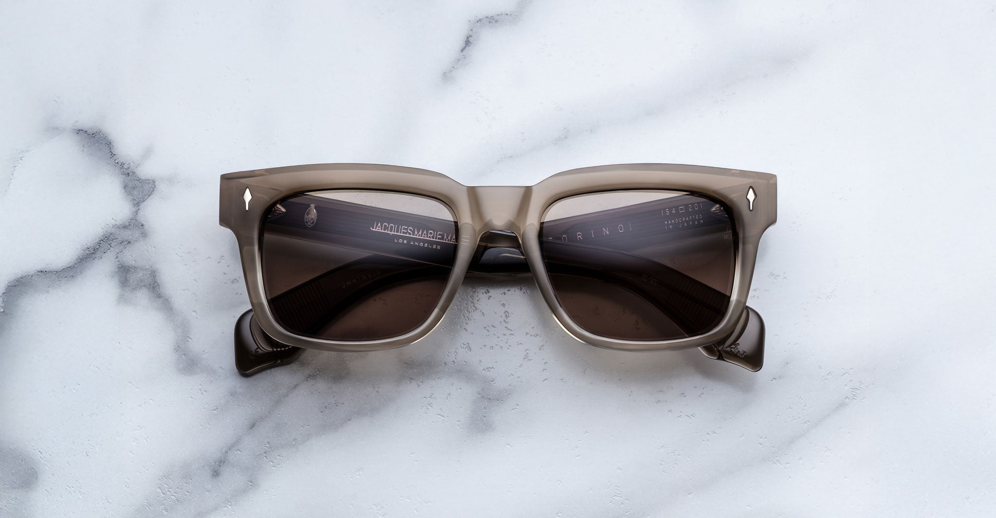 Jacques Marie Mage Torino sunglasses by Jacques Marie Mage, featuring square gray frames and dark lenses, are premium Japanese eyewear. Displayed on a white marble surface, they are photographed from above.