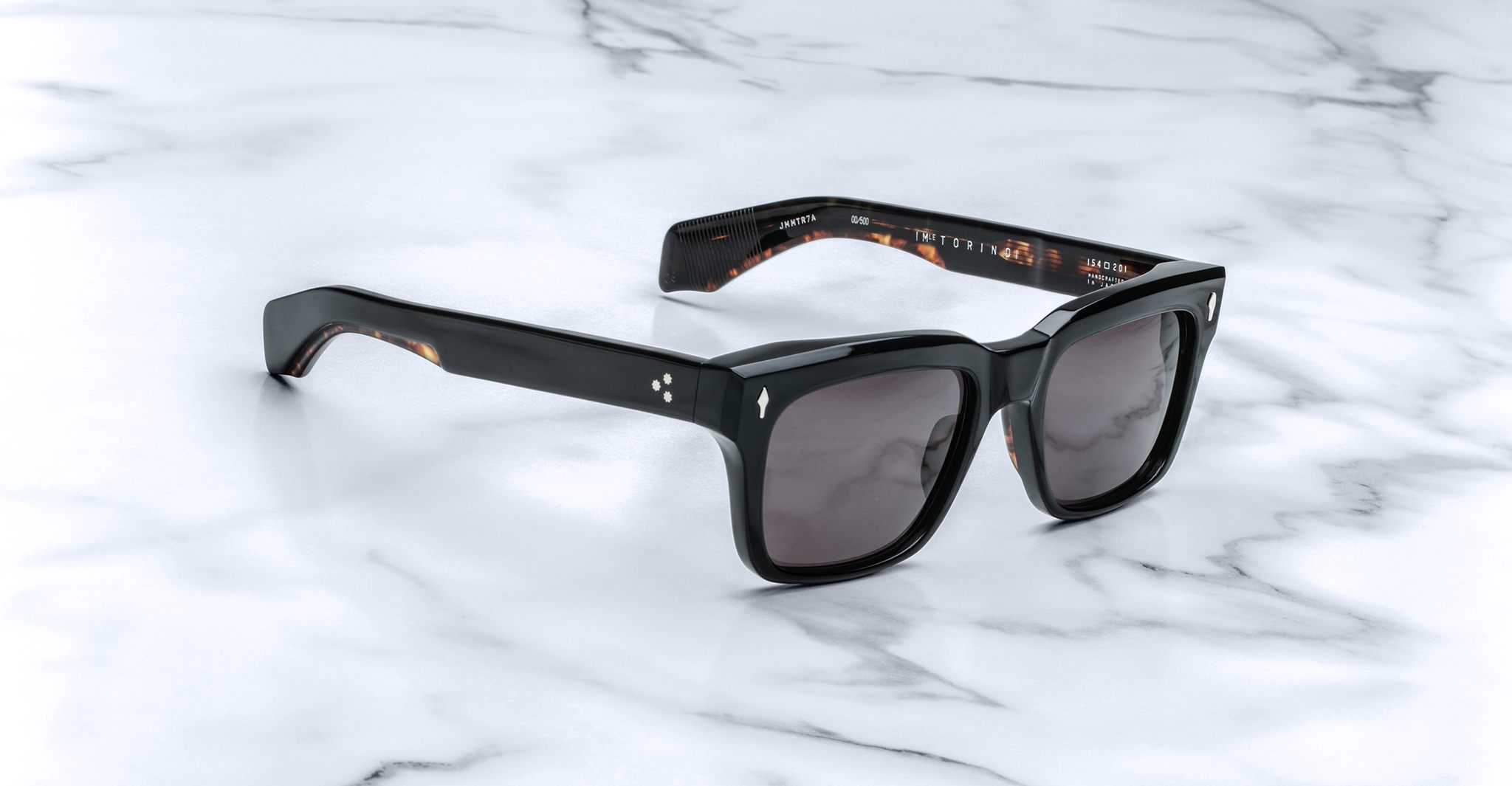 The Jacques Marie Mage Torino sunglasses feature bold black rectangular acetate frames and dark lenses, displayed on a white marble surface to highlight the sophisticated, handcrafted design of Jacques Marie Mage eyewear.