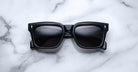 Jacques Marie Mage Torino sunglasses by Jacques Marie Mage feature bold black rectangular acetate frames and dark lenses, displayed on a white marble surface, recalling the handcrafted elegance of the Torino style.