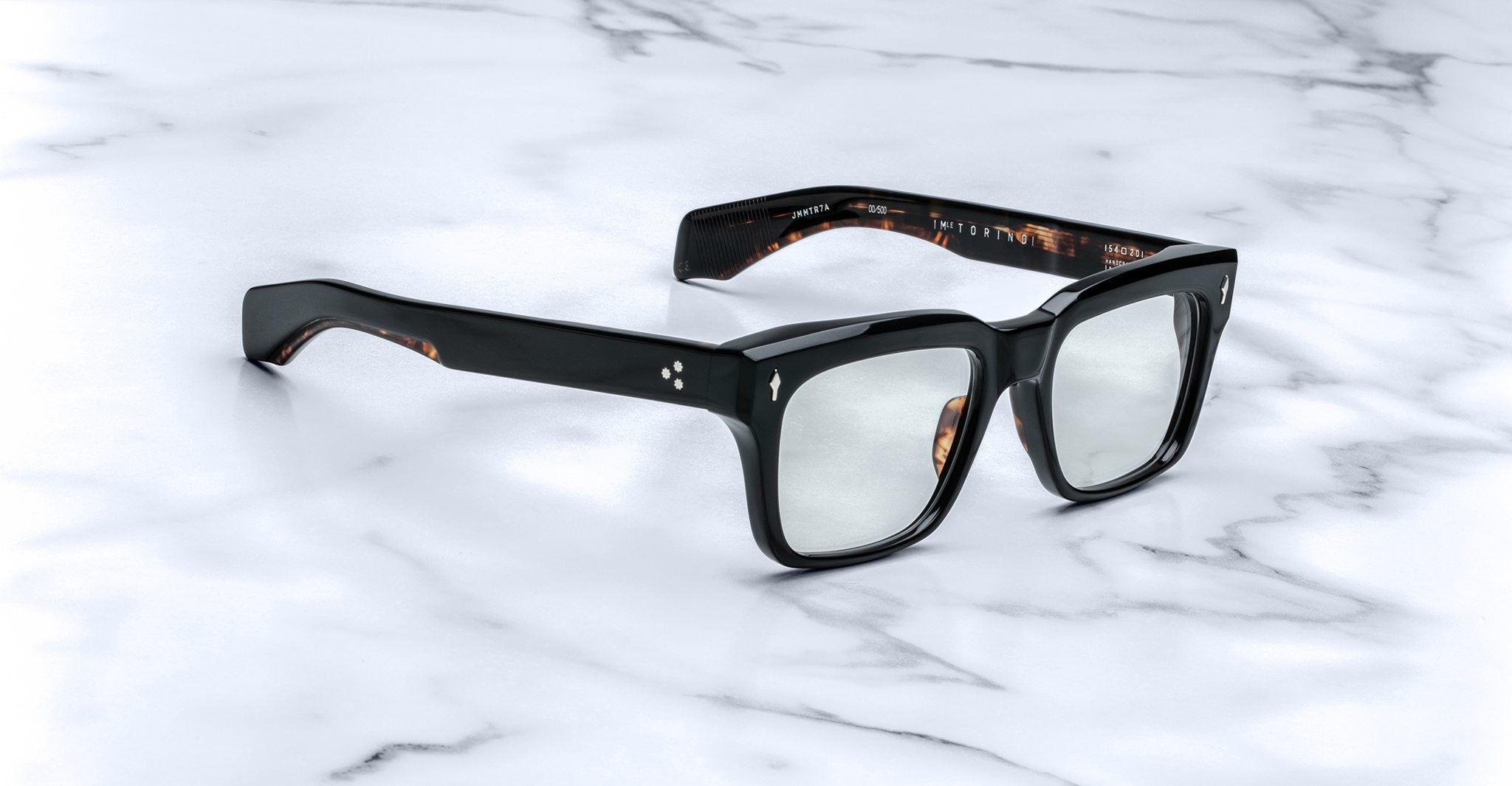 A pair of Jacques Marie Mage Torino black rectangular eyeglass frames by Jacques Marie Mage rests on a white marble surface.