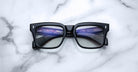 Jacques Marie Mage Torino eyeglasses feature bold black rectangular acetate frames with blue-tinted lenses, shown on a white marble surface. The digital screen reflection highlights the handcrafted design by Jacques Marie Mage.