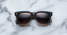 Jacques Marie Mage Torino sunglasses feature bold black square acetate frames with brown-tinted lenses, displayed on white marble—a striking example of handcrafted eyewear from Jacques Marie Mage.