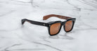 The Jacques Marie Mage Torino features bold black rectangular acetate frames, brown tinted lenses, and wood-textured inner arms, showcased on a white marble surface.