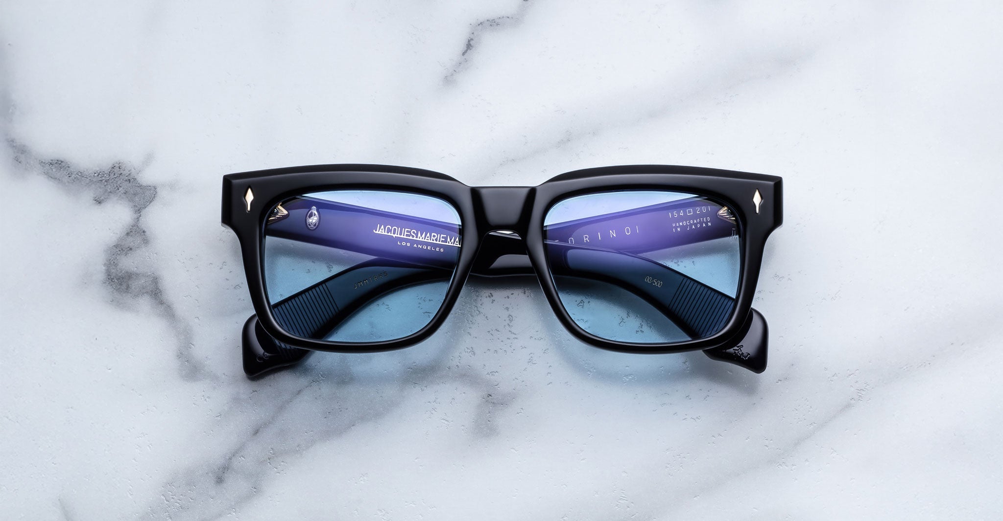 A pair of Jacques Marie Mage Torino rectangular frames with blue-tinted lenses sits on white marble, highlighting the refined craftsmanship of Jacques Marie Mage eyewear.