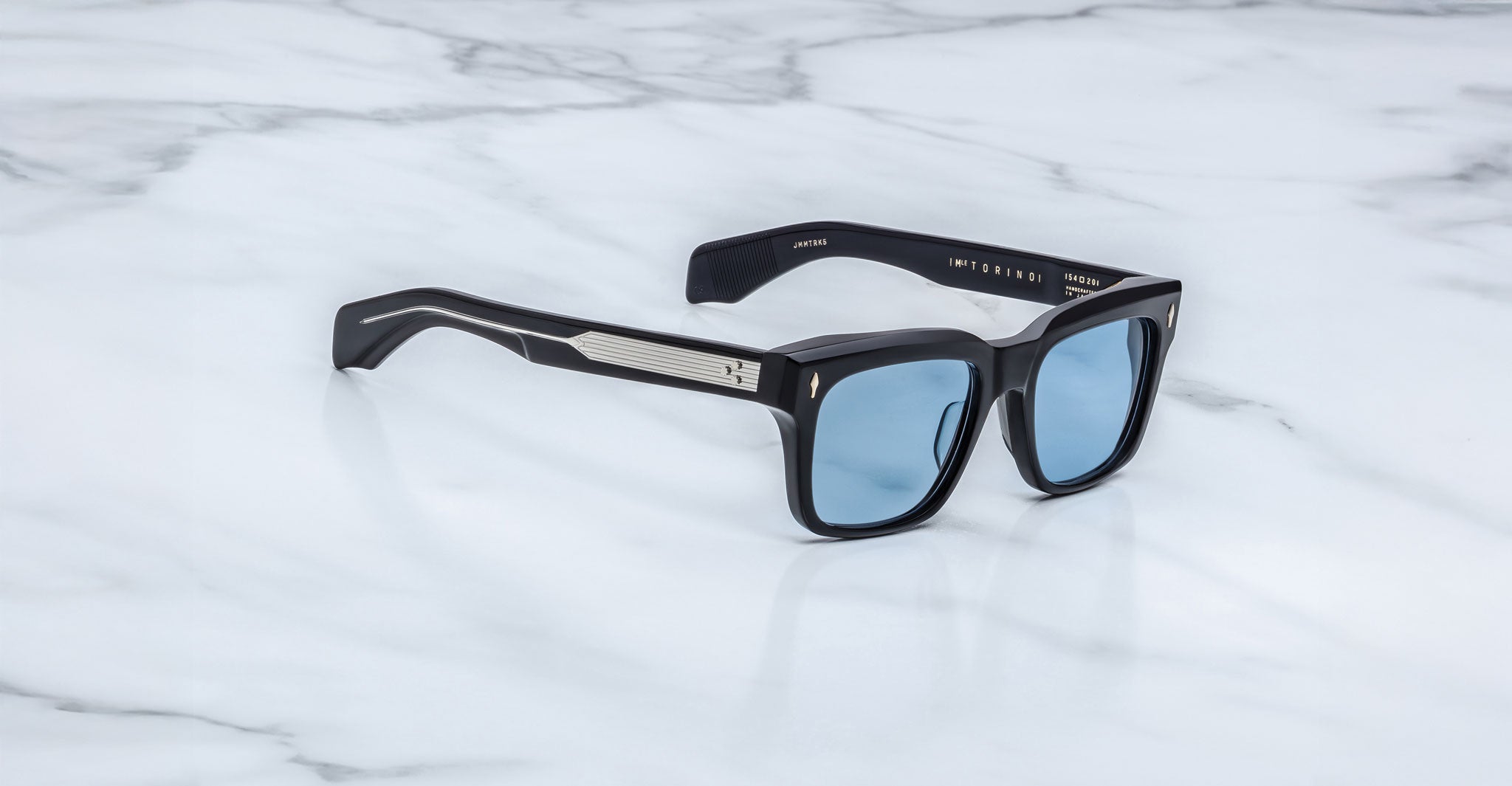 The Jacques Marie Mage Torino sunglasses by Jacques Marie Mage feature bold black rectangular acetate frames, blue-tinted lenses, and silver arm detailing, elegantly displayed on a white marble surface.