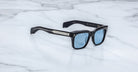 The Jacques Marie Mage Torino sunglasses by Jacques Marie Mage feature bold black rectangular acetate frames, blue-tinted lenses, and silver arm detailing, elegantly displayed on a white marble surface.