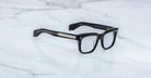 Jacques Marie Mage Torino eyeglasses by Jacques Marie Mage feature bold black rectangular acetate frames with silver arm detailing, elegantly displayed on a white marble surface.