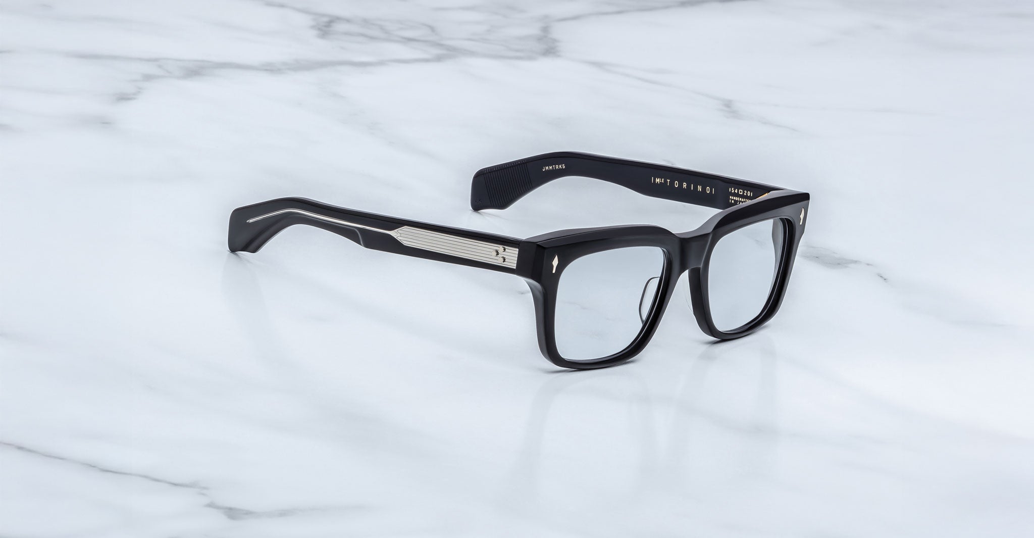 Jacques Marie Mage Torino eyeglasses by Jacques Marie Mage feature bold black rectangular acetate frames with silver arm detailing, elegantly displayed on a white marble surface.