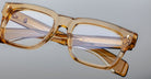 The Jacques Marie Mage Torino, bold rectangular eyeglass frames in amber transparent acetate by Jacques Marie Mage, rest on a light gray surface—a striking example of handcrafted acetate eyewear.