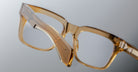 Close-up of Jacques Marie Mage Torino eyeglasses, featuring bold, rectangular frames in light brown translucent handcrafted acetate with clear lenses, shown against a neutral background.