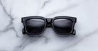 Jacques Marie Mage Torino sunglasses by Jacques Marie Mage, featuring bold black rectangular acetate frames with dark lenses, are displayed on a white marble surface.