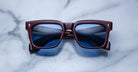The Jacques Marie Mage Torino sunglasses by Jacques Marie Mage feature bold square frames in dark red acetate with blue lenses, elegantly displayed on a white marble surface.