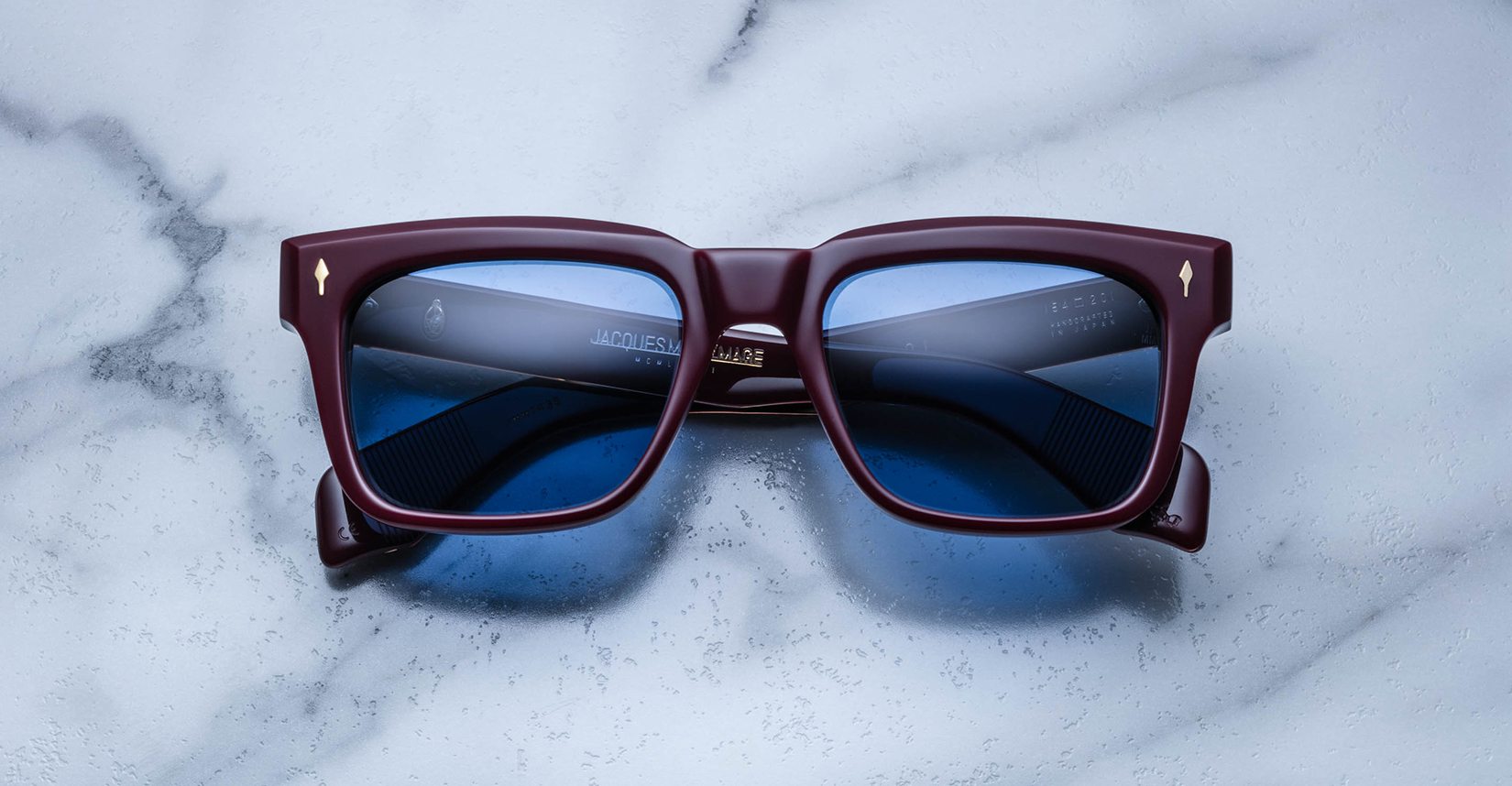 The Jacques Marie Mage Torino sunglasses by Jacques Marie Mage feature bold square frames in dark red acetate with blue lenses, elegantly displayed on a white marble surface.