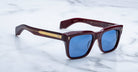 A pair of Jacques Marie Mage Torino square burgundy sunglasses with blue lenses and gold temple detailing—bold frames by Jacques Marie Mage—rest on a white marble surface.