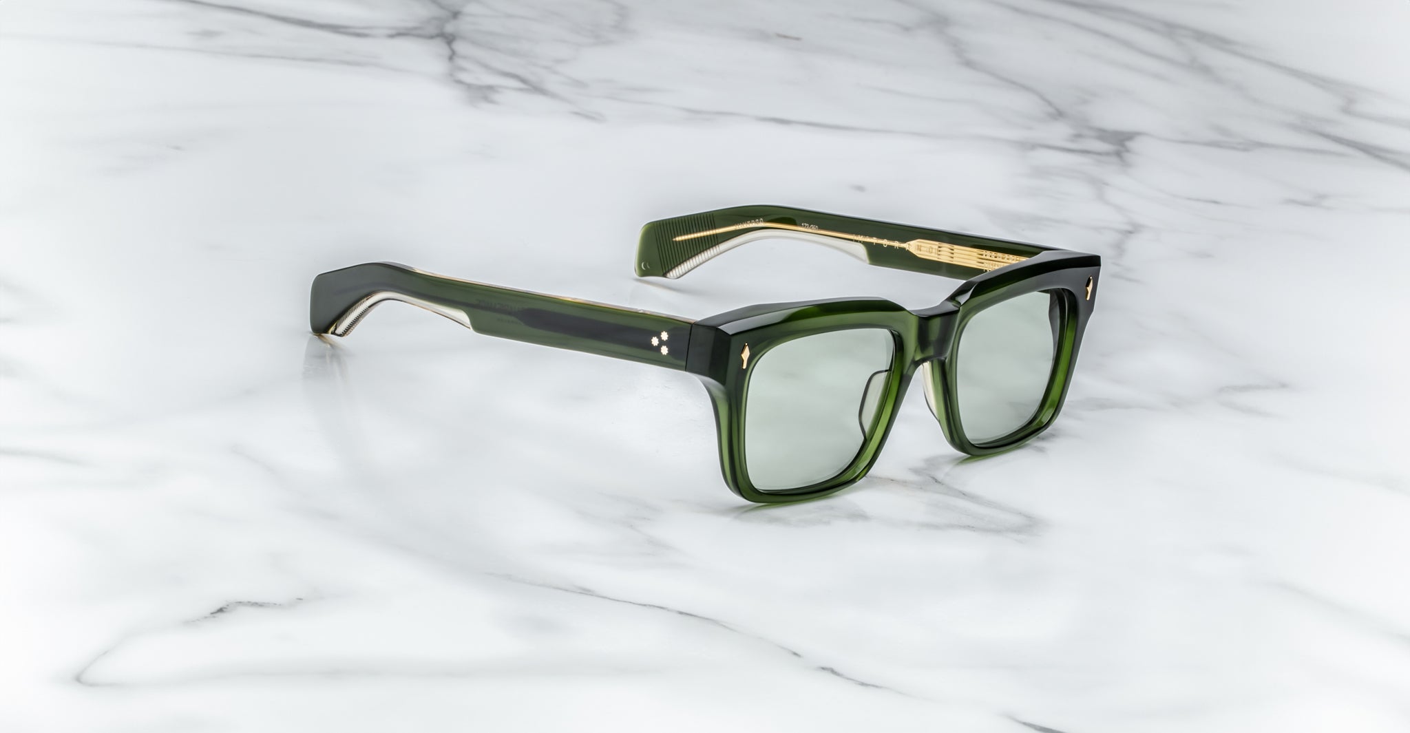 Jacques Marie Mage Torino sunglasses by Jacques Marie Mage, featuring a bold green rectangular design with gold accents, are displayed on a white marble surface.