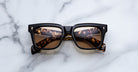 A pair of Jacques Marie Mage Torino rectangular sunglasses, featuring handcrafted acetate frames in tortoiseshell and black with amber lenses, rest on a white marble surface.