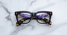 Jacques Marie Mage Torino eyeglasses feature bold, handcrafted black acetate temples and rectangular frames. Resting on marble, their lenses reflect city lights—showcasing the iconic style of Jacques Marie Mage.