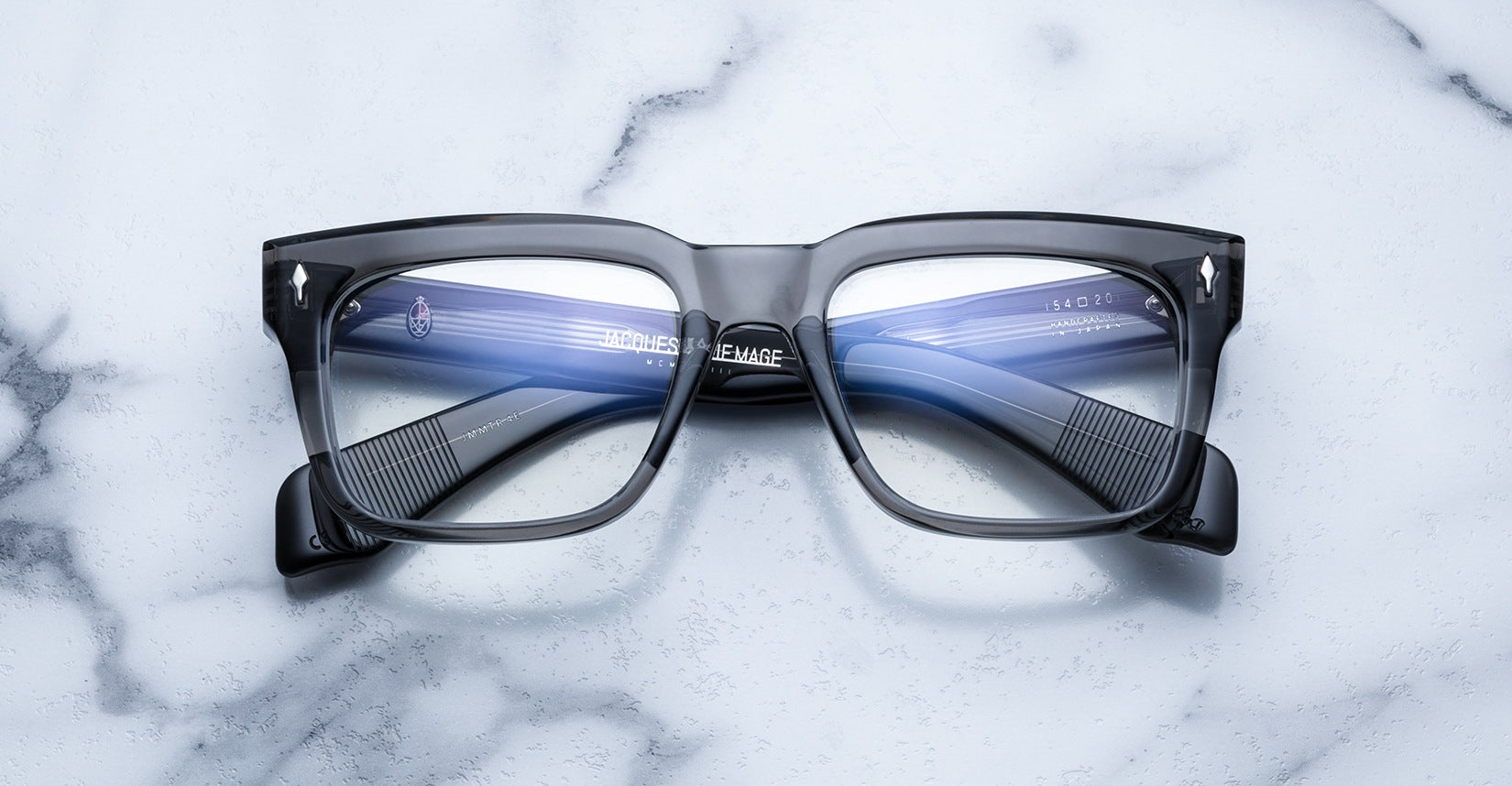 A pair of Jacques Marie Mage Torino rectangular eyeglass frames by Jacques Marie Mage, featuring blue-tinted lenses and handcrafted from premium acetate, displayed on a white marble surface.