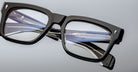 Close-up of Jacques Marie Mage Torino bold rectangular eyeglass frames, with a blue and silver fountain pen reflected in the lenses against a light gray background—showcasing the refined, handcrafted acetate eyewear by Jacques Marie Mage.