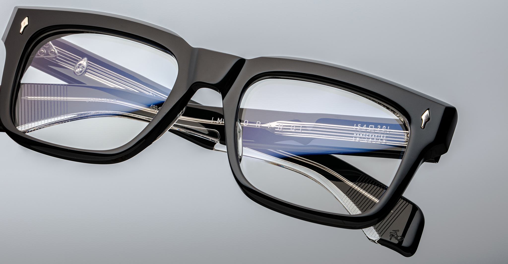 Close-up of Jacques Marie Mage Torino bold rectangular eyeglass frames, with a blue and silver fountain pen reflected in the lenses against a light gray background—showcasing the refined, handcrafted acetate eyewear by Jacques Marie Mage.