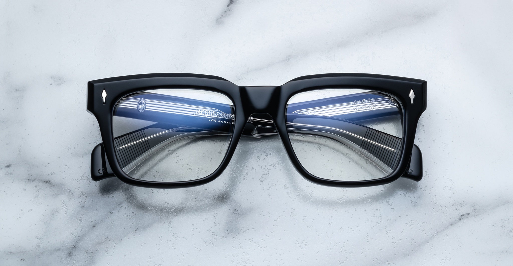 The Jacques Marie Mage Torino eyeglass frames by Jacques Marie Mage feature bold rectangular design with striped blue and white reflections on the lenses, displayed on a white marble surface.