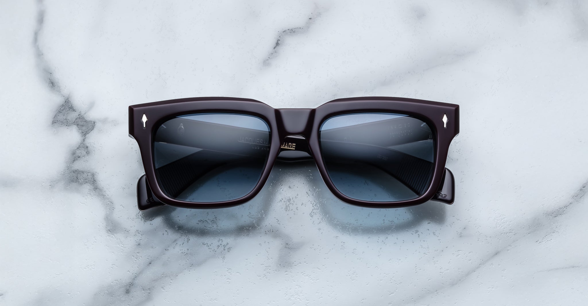 The Jacques Marie Mage Torino sunglasses by Jacques Marie Mage feature bold black rectangular frames and dark lenses, elegantly displayed on a white marble surface.