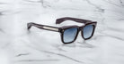The Jacques Marie Mage Torino by Jacques Marie Mage features black handcrafted acetate rectangular frames, blue gradient lenses, and metallic arm accents, shown on a white marble surface.