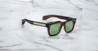 These bold square brown Jacques Marie Mage Torino sunglasses feature green lenses and metallic temple accents, showcasing handcrafted acetate frames displayed elegantly on a white marble surface.