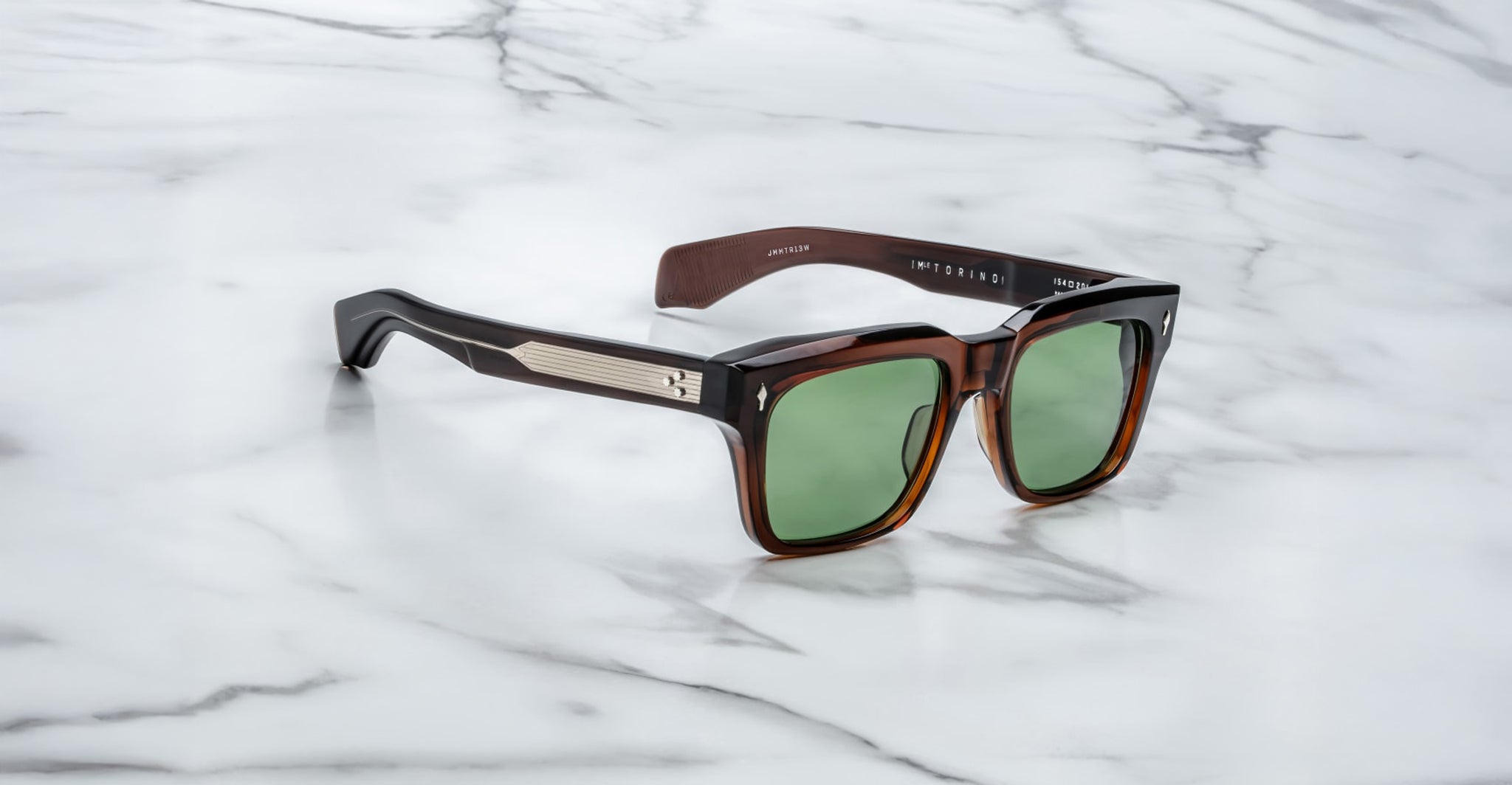 These bold square brown Jacques Marie Mage Torino sunglasses feature green lenses and metallic temple accents, showcasing handcrafted acetate frames displayed elegantly on a white marble surface.