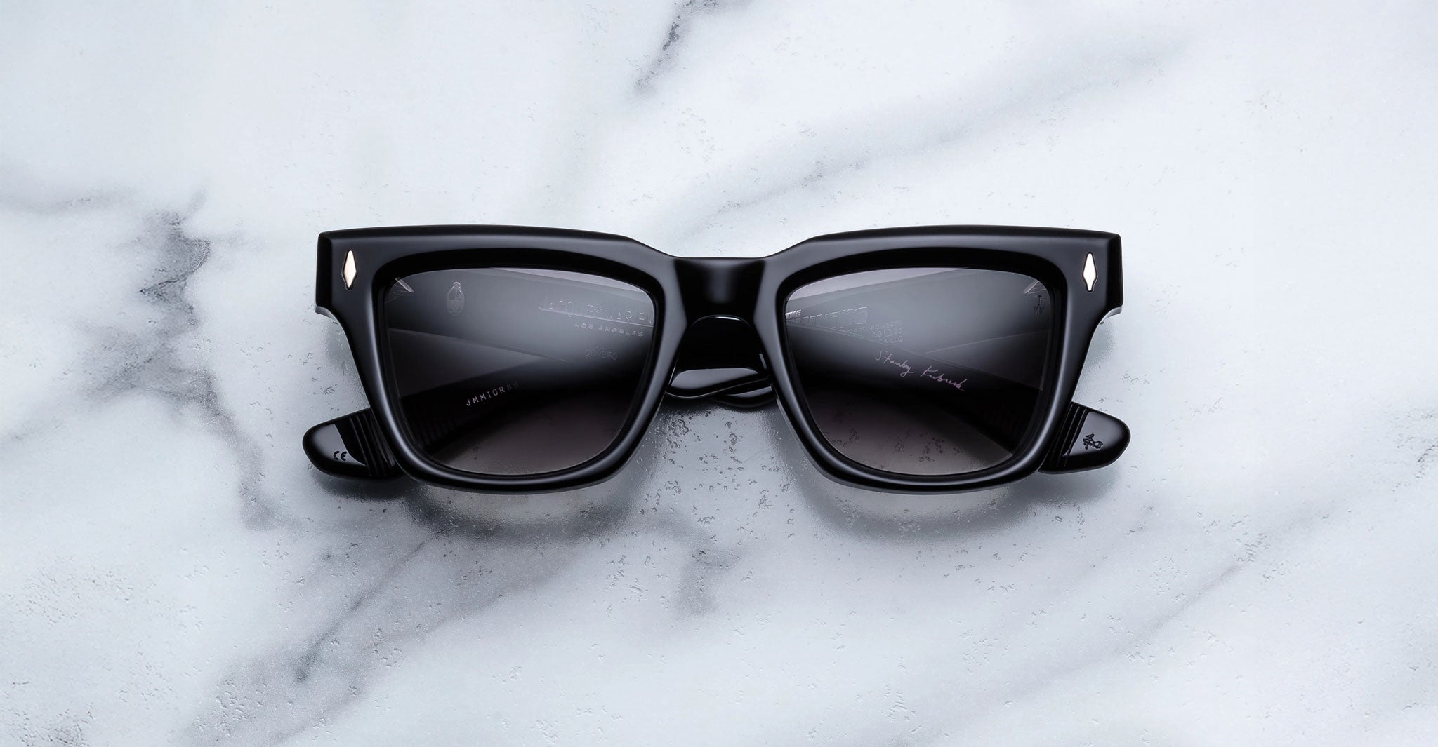 A pair of Jacques Marie Mage Torrance black square sunglasses with dark lenses from the limited-edition rectangular frame collection rests on a white marble surface.