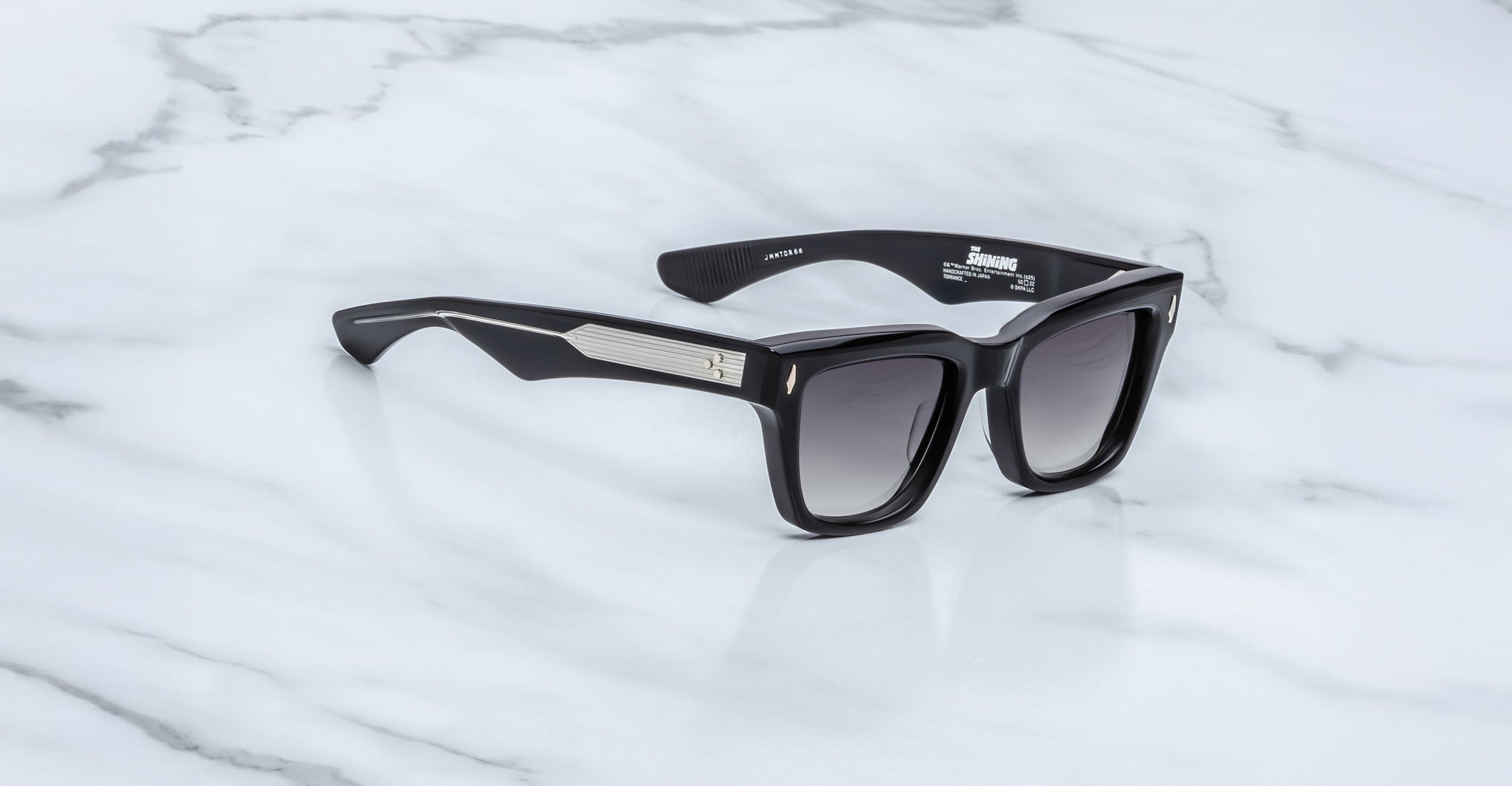 The Jacques Marie Mage Torrance sunglasses feature a black rectangular frame from The Shining Collection with dark gradient lenses and silver arm accents—a limited-edition style showcased on white marble.