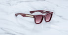 A pair of glossy maroon Jacques Marie Mage Torrance sunglasses with gradient lenses and gold temple detailing, from the limited-edition rectangular The Shining Collection, displayed on a white marble surface.