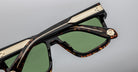 Close-up of tortoiseshell sunglasses with green lenses, gold accents along the top frame, and a decorative gold emblem near the hinge, set against a plain gray background.