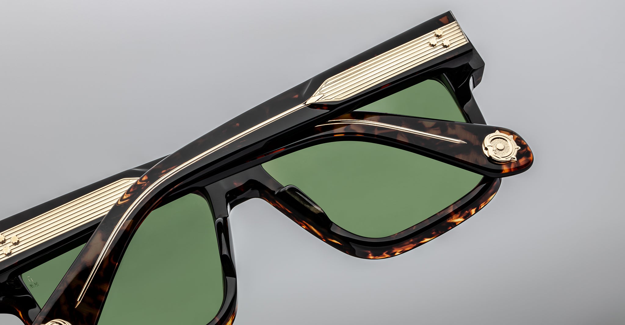 Close-up of tortoiseshell sunglasses with green lenses, gold accents along the top frame, and a decorative gold emblem near the hinge, set against a plain gray background.