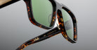 Close-up of tortoiseshell sunglasses with green lenses, showing detailed frame texture and the inscription IME TRAMONTO I in gold on the arm. The background is a smooth, neutral gray.
