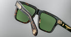 Close-up of stylish tortoiseshell sunglasses with green lenses, gold detailing on the arms, and a gold emblem on the hinge, set against a plain, light gray background.