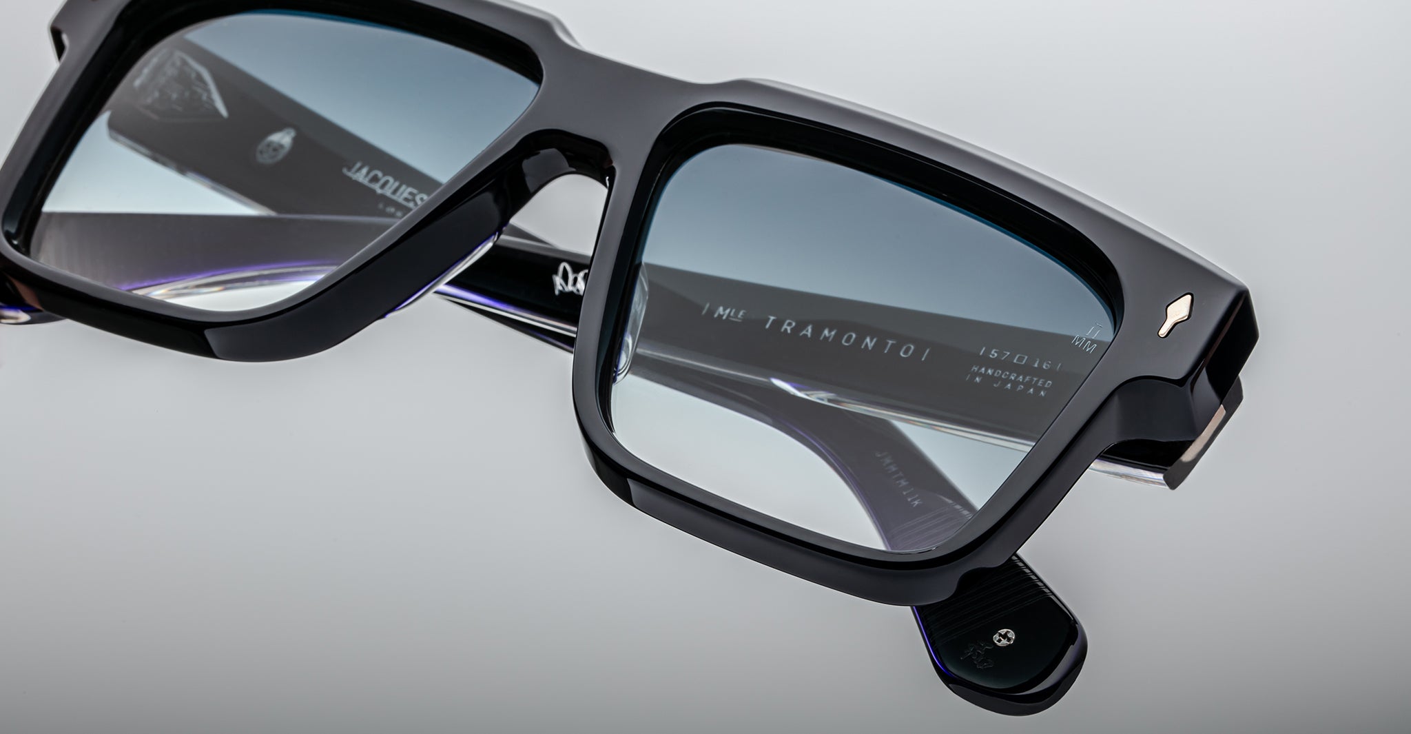 Close-up view of stylish black eyeglasses with rectangular frames, featuring the brand name TRAMONTO and other text on the inside of the arm, displayed on a reflective, light gray surface.