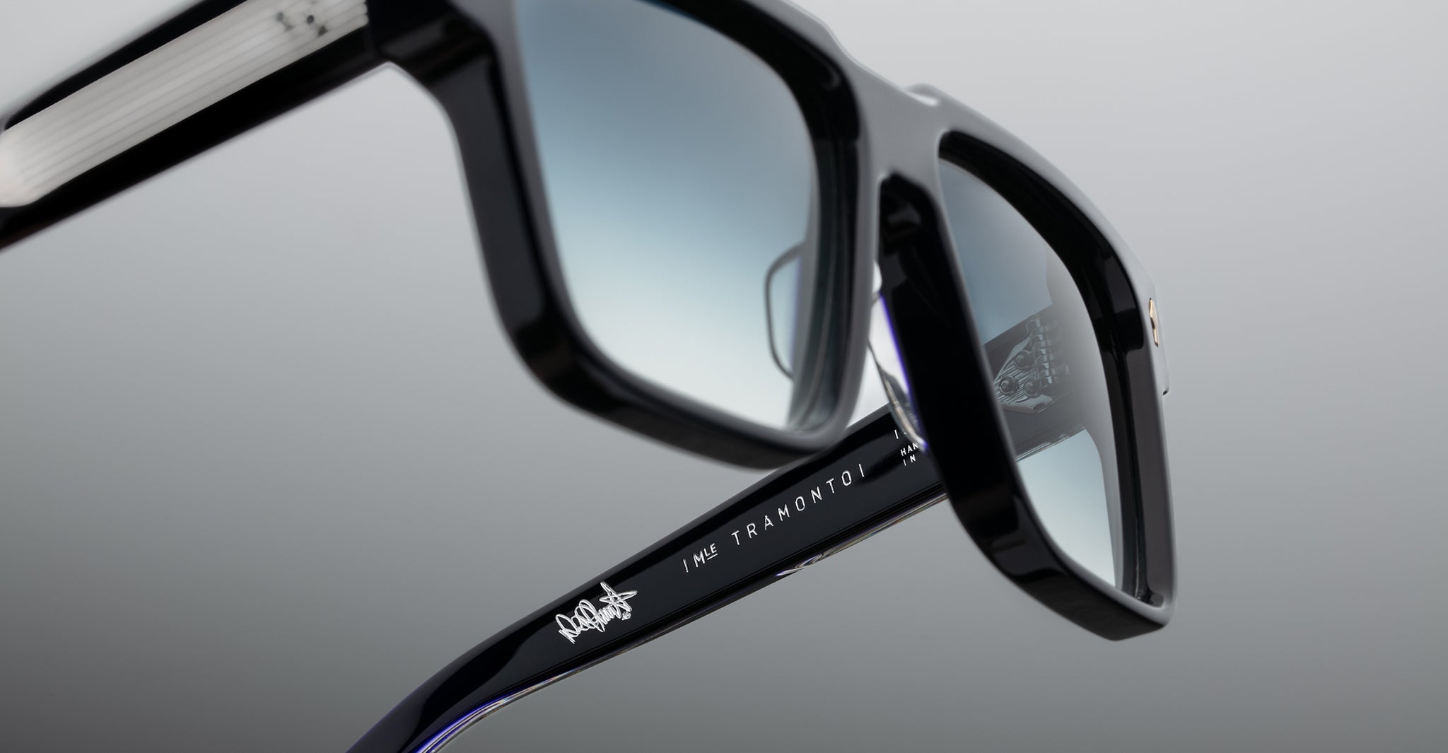 Close-up of black-framed sunglasses with gradient lenses, showing the engraved text “IME TRAMONTONI” and a small white logo on the arm, against a plain, blurred background.