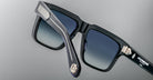 A close-up of stylish black square sunglasses with blue gradient lenses, showing detailed metallic embellishments on the hinge and temple tips, set against a neutral gray background.